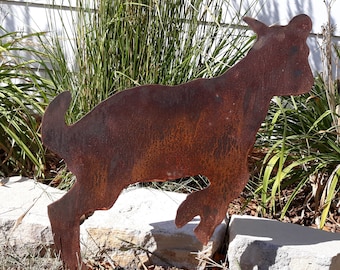 May include: A rusty metal silhouette of a goat, standing on a bed of green grass and rocks. The goat is facing right and has a long, thin tail.