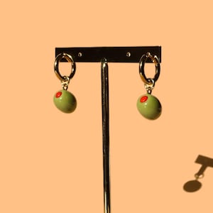 May include: A pair of gold hoop earrings with green olive-shaped charms. The earrings are displayed on a wooden stand.