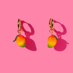 May include: A pair of gold hoop earrings, each with a mango charm. The mangoes are yellow, orange, and green, with a small green leaf. The earrings are set against a bright pink background.