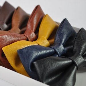 May include: A collection of eight bow ties made from textured faux leather in various colors including black, navy blue, mustard yellow, brown, reddish-brown, dark burgundy, and dark olive green, arranged in a row within a white rectangular tray.