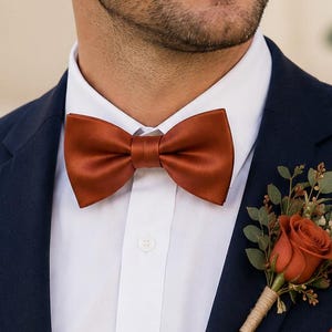 May include: A navy blue suit jacket worn with a crisp white button-down shirt, a solid burnt orange satin bow tie, a matching burnt orange pocket square, and a floral boutonniere featuring a single burnt orange rose with green foliage.