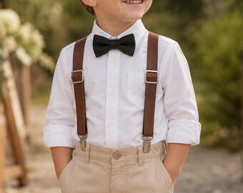 May include: A white long-sleeved button-down shirt paired with tan trousers, brown leather suspenders featuring silver-toned metal hardware, and a solid black bow tie. The shirt is tucked into the trousers, and the sleeves are rolled up.