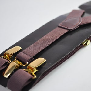 Puede incluir: Un par de tirantes marrones con clips de metal dorado. Los tirantes están empaquetados en un fondo negro con una cremallera.