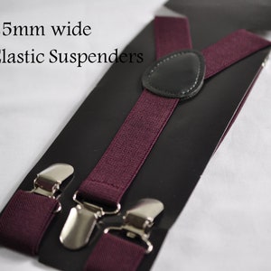 May include: Deep burgundy elastic suspenders featuring a 25mm wide band, silver-toned metal clips, and a central black leather-like patch. The product is displayed on a black backing card with the text 25mm wide Elastic Suspenders printed on it.