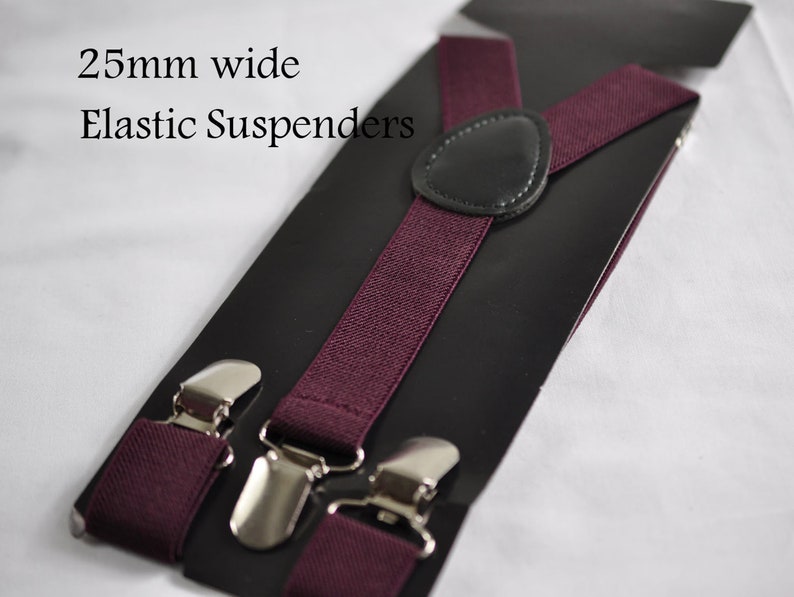 May include: Deep burgundy elastic suspenders featuring a 25mm wide band, silver-toned metal clips, and a central black leather-like patch. The product is displayed on a black backing card with the text 25mm wide Elastic Suspenders printed on it.