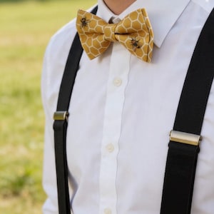 May include: A yellow bow tie featuring a white honeycomb pattern and small bee illustrations, worn with a crisp white button-down shirt and black braces that have metallic gold-toned hardware clips attached to the fabric straps.