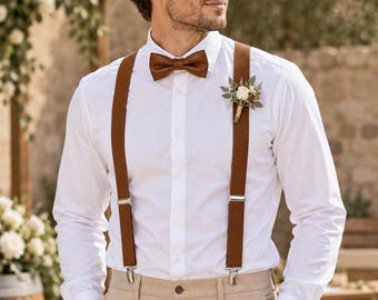 May include: A crisp white long-sleeved button-down shirt paired with tan trousers, featuring brown suspenders and a matching brown bow tie. A small floral boutonniere with a white rose and green foliage is pinned to the left side of the shirt.