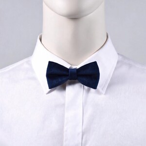 May include: A navy blue velvet bow tie is centered on a white collared shirt. The bow tie is a classic accessory, adding a touch of elegance. The fabric has a soft, textured appearance. The bow tie is a dark, solid color.