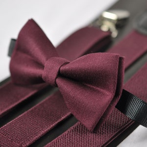 May include: A deep burgundy bow tie with a textured fabric finish, paired with matching burgundy elastic suspenders featuring black ribbon accents and silver-toned metal clips for secure attachment to clothing items.