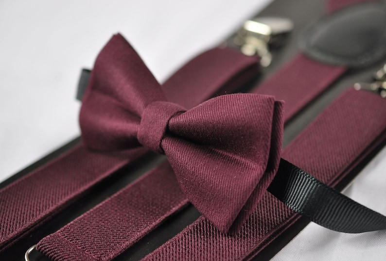 May include: A deep burgundy bow tie with a textured fabric finish, paired with matching burgundy elastic suspenders featuring black ribbon accents and silver-toned metal clips for secure attachment to clothing items.