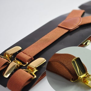 May include: Brown suspenders with gold metal clips. The suspenders are made of a soft, woven fabric and have a leather patch on one strap.