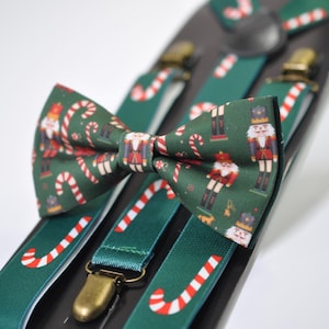 May include: A green bow tie and suspenders set with a Christmas theme. The bow tie and suspenders feature a repeating pattern of candy canes and nutcrackers. The suspenders have gold-coloured metal clips.