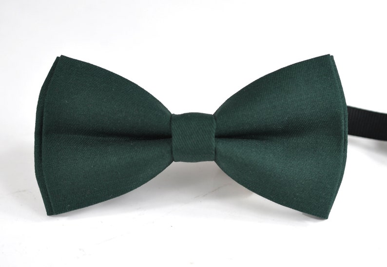 May include: A solid dark forest green bow tie featuring a classic pre-tied shape with a central knot and a smooth, matte fabric texture. The bow tie includes a thin, black adjustable strap extending from the right side for secure attachment.