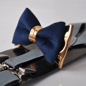 Puede incluir: Una pajarita azul marino con un cierre de metal dorado y un par de tirantes grises con clips de metal plateado.