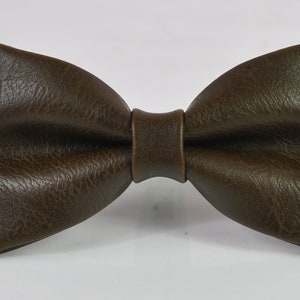 May include: A dark brown bow tie with a textured, leather-like surface. The bow tie features a classic pre-tied shape with a central knot detail, showcasing a consistent deep brown color and a visible grain pattern across the entire surface.