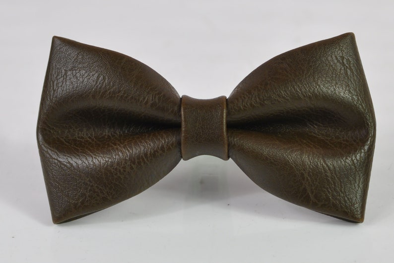May include: A dark brown bow tie with a textured, leather-like surface. The bow tie features a classic pre-tied shape with a central knot detail, showcasing a consistent deep brown color and a visible grain pattern across the entire surface.