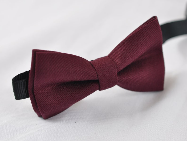 May include: A deep burgundy bow tie featuring a textured fabric weave. The bow tie has a central knot and is attached to a black adjustable strap that extends from the sides, showcasing a classic pre-tied shape with clean, structured edges.