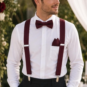 May include: A crisp white long-sleeved button-down shirt paired with deep burgundy braces, a matching burgundy bow tie, and a burgundy pocket square. The braces feature silver-toned metal clips and adjusters, worn over black trousers.