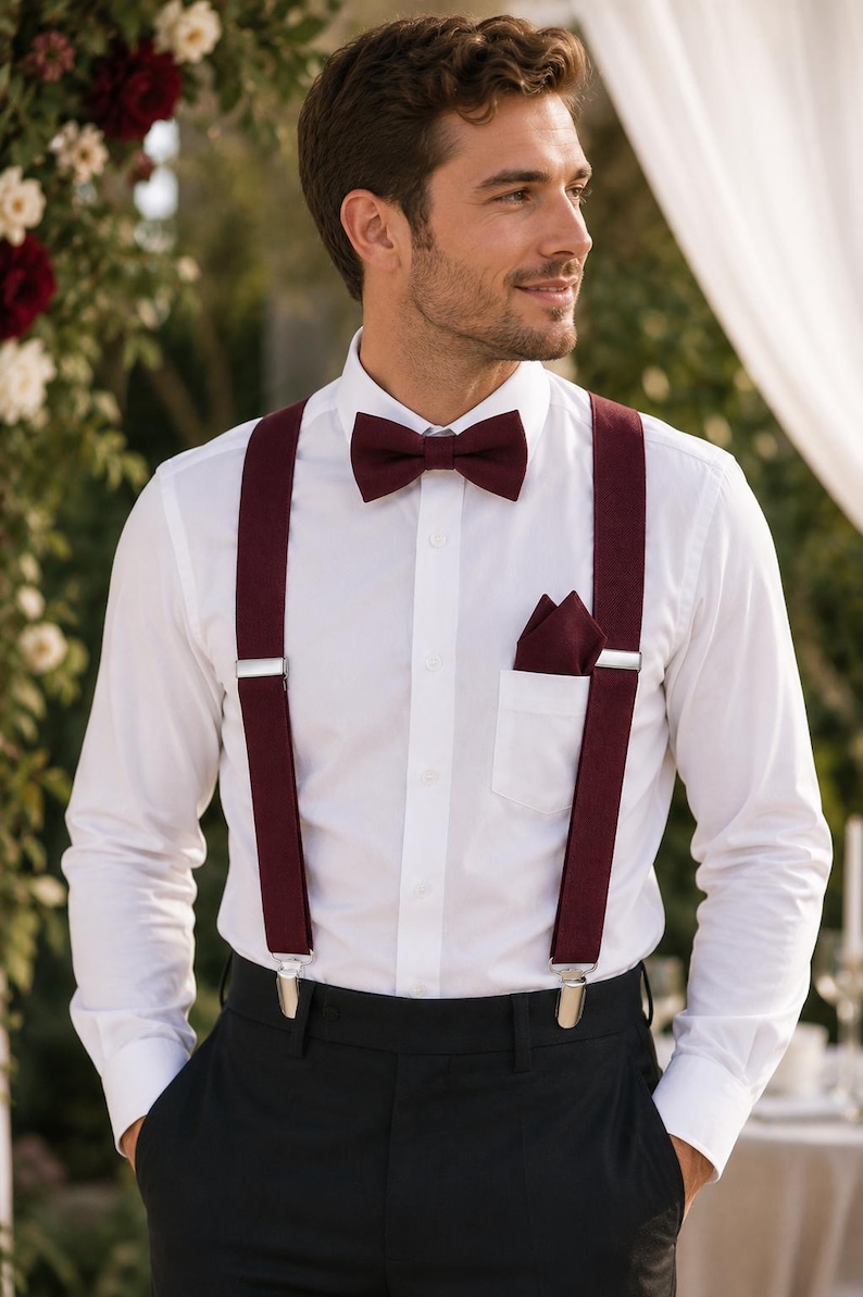 May include: A crisp white long-sleeved button-down shirt paired with deep burgundy suspenders, a matching burgundy bow tie, and a burgundy pocket square. The suspenders feature silver-toned metal clips and adjusters, worn over black trousers.