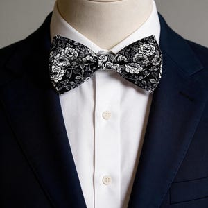 May include: A black bow tie featuring a white floral pattern with detailed rose illustrations. The bow tie is worn over a crisp white button-down shirt and a dark navy blue blazer, creating a sharp contrast between the dark fabric and the white print.