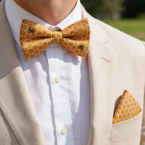 Può includere: Un papillon giallo senape e una pochette abbinata con un motivo esagonale a nido d'ape bianco e piccole illustrazioni di api. Il set è indossato con una camicia bianca e un blazer beige chiaro, creando un look coordinato ed elegante.