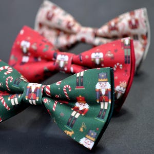 May include: Three festive bow ties with holiday-themed patterns. One is green with candy canes and nutcrackers, another is red with nutcrackers, and the third is white with holiday motifs. Perfect for adding a touch of holiday cheer to any outfit.