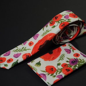 May include: A white tie and pocket square set featuring a vibrant floral pattern of red poppies, purple flowers, and green leaves. The tie is partially rolled, and the pocket square is folded, both on a dark background.