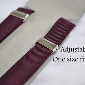 May include: A pair of burgundy elastic suspenders featuring polished silver-tone metal clips for adjustment. The product is displayed on a light gray card with the printed text: Adjustable, One size fits most. The fabric has a woven texture.