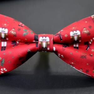 Könnte beinhalten: Eine rote Fliege mit einem sich wiederholenden Muster aus Nussknackern, Weihnachtsbäumen und Zuckerstangen. Dieses festliche Accessoire ist ideal für Weihnachtsfeiern. Der Stoff hat einen leichten Glanz.
