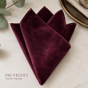 May include: A deep burgundy velvet pocket square folded into a multi-point shape. The fabric has a soft, matte texture and a rich, dark red color. The text FIG VELVET POCKET SQUARE is printed in the lower left corner of the image in a dark font.