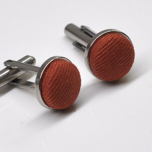 Op de afbeelding: Een paar manchetknopen met een zilveren metalen basis en een ronde, oranje stoffen bovenkant.