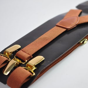 May include: Brown suspenders with gold metal clips. The suspenders are packaged on a black backing with a brown zipper.