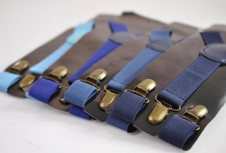 Puede incluir: Un juego de seis tirantes en varios tonos de azul con clips de metal dorado antiguo. Los tirantes est&aacute;n dispuestos en una fila sobre un fondo negro.