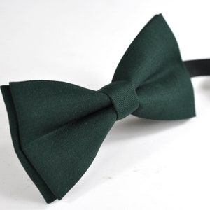May include: A dark forest green bow tie featuring a classic pre-tied shape with a central fabric knot. The bow tie is constructed from a solid-coloured textile material and includes a thin black adjustable strap extending from the right side.