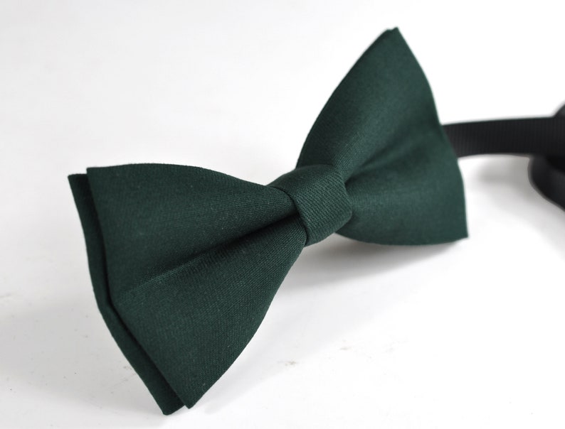 May include: A dark forest green bow tie featuring a classic pre-tied shape with a central fabric knot. The bow tie is constructed from a solid-coloured textile material and includes a thin black adjustable strap extending from the right side.