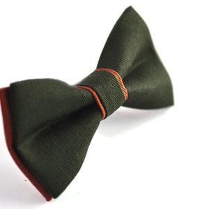 May include: A dark green bow tie with a rust-colored border and accents. The bow tie is made of a textured fabric and is set against a white background. This formal accessory is suitable for special occasions.