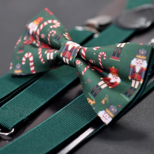 May include: A festive green bow tie and suspenders set. The bow tie features a pattern of nutcrackers, candy canes, and Santa Claus figures. The suspenders are a solid dark green with silver-colored metal clips.