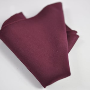 May include: A solid deep burgundy pocket square made of a smooth, matte fabric. The square is folded into a triangular shape, showcasing its rich, dark red color and clean, straight edges. The texture appears uniform and consistent throughout.