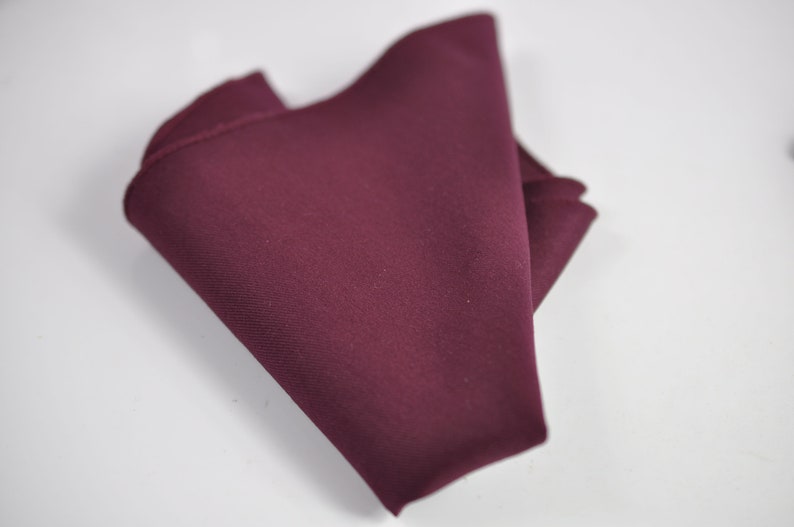 May include: A solid deep burgundy pocket square made of a smooth, matte fabric. The square is folded into a triangular shape, showcasing its rich, dark red color and clean, straight edges. The texture appears uniform and consistent throughout.
