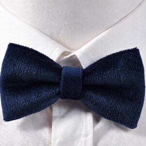 May include: A navy blue velvet bow tie is centered against a white dress shirt. The bow tie has a soft, textured appearance and is secured with a matching fabric band. The classic accessory adds a touch of elegance.