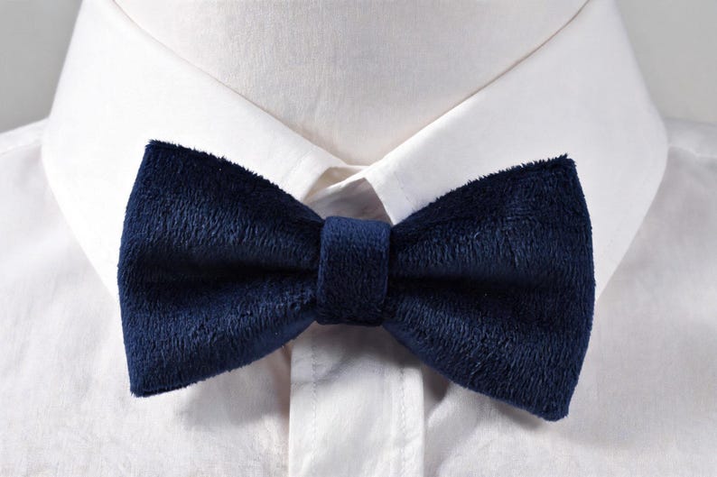 May include: A navy blue velvet bow tie is centered against a white dress shirt. The bow tie has a soft, textured appearance and is secured with a matching fabric band. The classic accessory adds a touch of elegance.