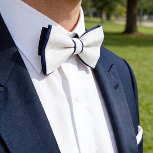 May include: A white bow tie with navy blue trim is the focal point of this image. The bow tie is set against a white dress shirt and a navy blue suit jacket. The image is a close-up, focusing on the upper chest and neck area.