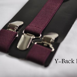 May include: Deep burgundy elastic suspenders featuring a Y-back design with polished silver-tone metal clips. The fabric has a woven texture and is attached to a black display card. The text Y-Back Design is printed in the bottom right corner.