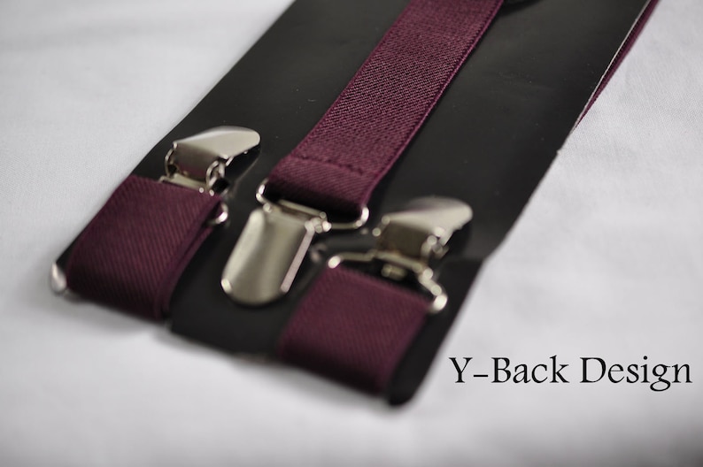 May include: Deep burgundy elastic suspenders featuring a Y-back design with polished silver-tone metal clips. The fabric has a woven texture and is attached to a black display card. The text Y-Back Design is printed in the bottom right corner.