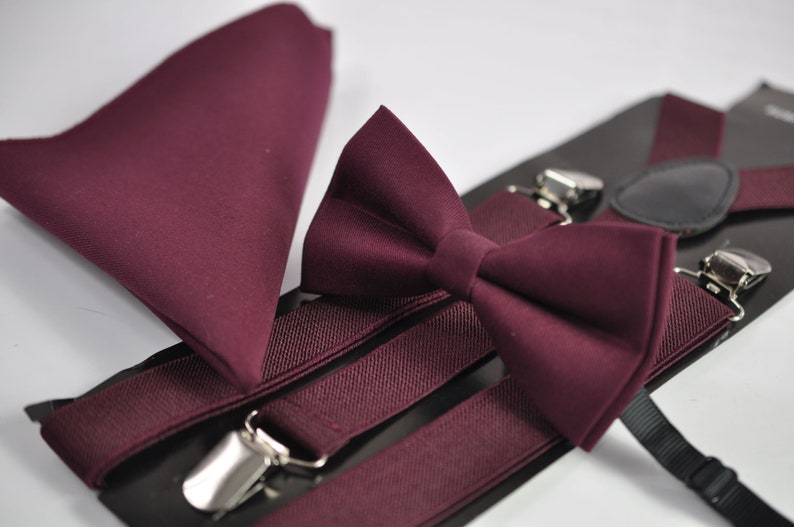 May include: A set consisting of a solid burgundy bow tie, a matching pocket square, and elastic suspenders with silver-toned metal clips. The items are presented on a black display card, showcasing a uniform deep red fabric texture throughout.