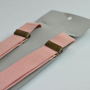 Peut inclure: Une paire de bretelles roses avec des clips en laiton. Les bretelles sont pli&eacute;es en deux et sont sur un fond gris.