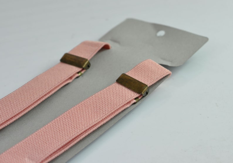 Peut inclure: Une paire de bretelles roses avec des clips en laiton. Les bretelles sont pli&eacute;es en deux et sont sur un fond gris.
