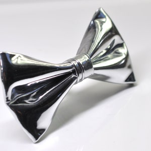 May include: A silver bow tie with a shiny, reflective surface. The bow tie is made of a smooth, pliable material and has a classic, elegant design.