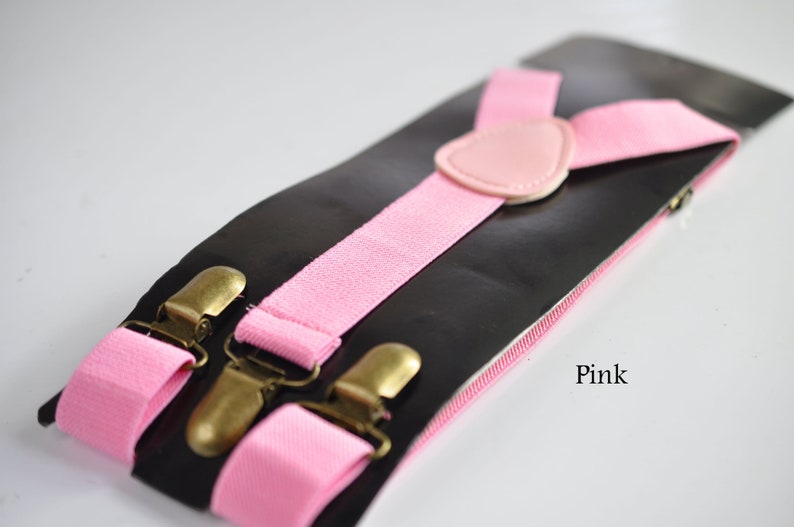 Peut inclure: Bretelles roses avec des clips dor&eacute;s et une sangle r&eacute;glable en cuir rose. Les bretelles sont sur un fond noir avec le mot "Rose" imprim&eacute; en bas &agrave; droite.