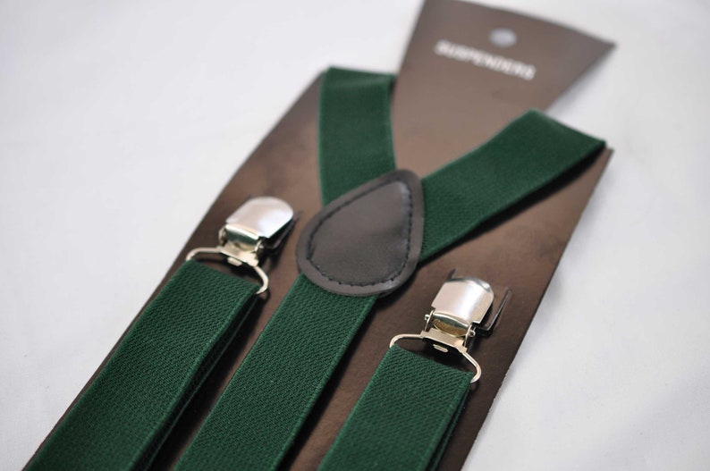 May include: Dark forest green elastic braces featuring silver-toned metal clips and a central dark brown faux leather patch. The braces are attached to a brown cardboard display card with the word SUSPENDERS printed in white text.